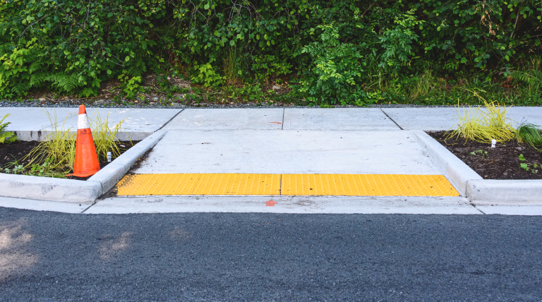 A sidewalk curb near a safety cone near and an ADA-compliant curb ramp