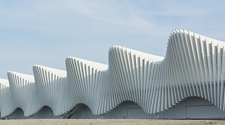 The side of a modern train station with walls in the shape of massive flowing white waves beneath a hazy blue sky