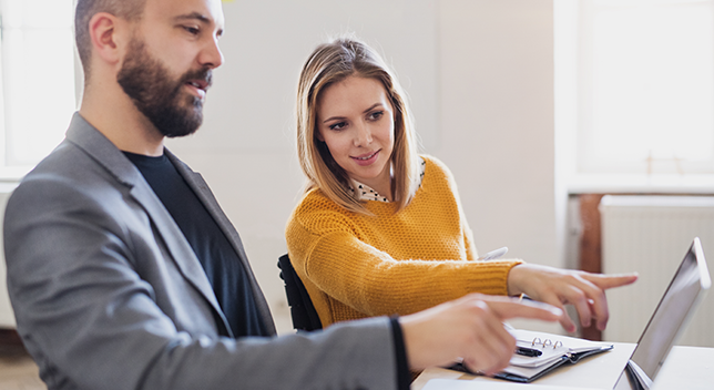 Two business professionals sit in an office setting, discussing something on a laptop which the viewer cannot see.