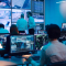 Multiple workers in a command center working on computers with multiple monitors displaying charts, text, and numerical data 