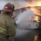 A burning home with bright orange flames and gray smoke and 3 firefighters in yellow uniforms holding a long yellow hose