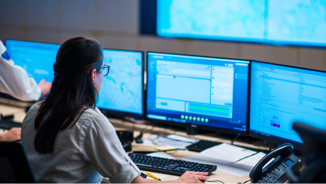 A woman at a command center viewing multiple computer screens and moving the black mouse 
