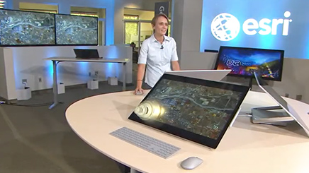 Blue background with the word Esri on it and a woman in front of a table and four large computer monitors