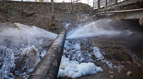 A large broken pipe, also known as a watermain, with water spraying out of it into a drainage ditch
