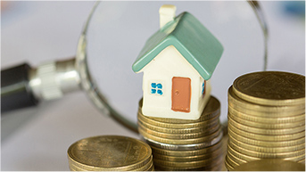 Small porcelain home on top of a stack of gold coins and a magnifying glass