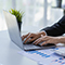 A person wearing a dark suit typing on a slim modern laptop on a minimalist white desk