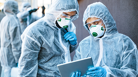 Two people wearing medical personal protective equipment and looking at a tablet together
