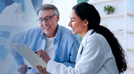 A doctor in a white lab coat smiling as she reviews a chart on a clipboard with a smiling patient wearing a blue collared shirt in a sunlit white office
