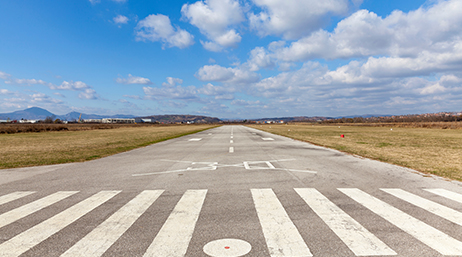An airplane runway stretching into the middle distance with brown and green fields on either side under a bright blue cloud-swept sky