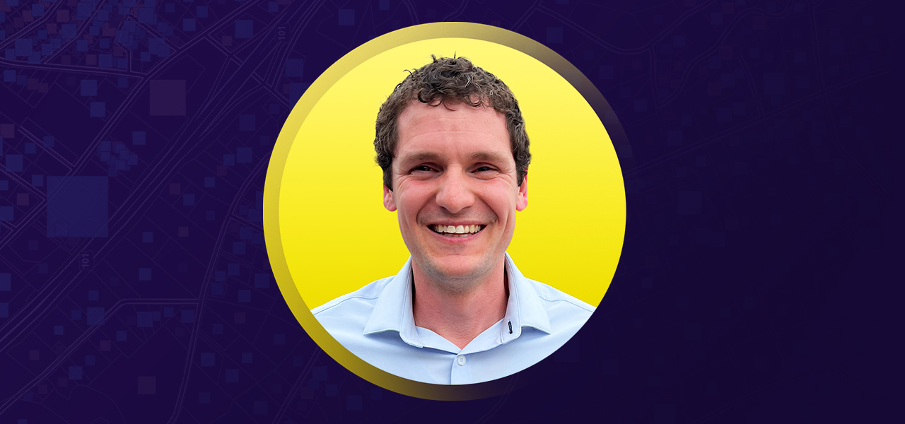 Portrait of Jonathan Blazs smiling in a light blue collared shirt with a flat yellow background in a circular border
