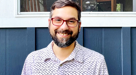 Anthony Pologruto with a short dark hair and a short beard, wearing red glasses and a white collared shirt with a grey diamond pattern