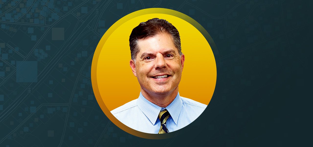 Portrait of Tony Garro smiling in a light blue collared shirt and tie with a flat yellow background in a round border