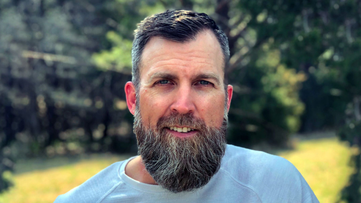 Portrait of Darin Welch smiling in a blue tee shirt with a tree-lined sunny field in the background