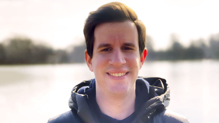 Portrait of George Floros smiling in a blue windbreaker with a tree-lined lake in the background