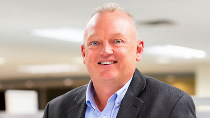 Portrait of Steve Anderson smiling in a gray coat and blue shirt with a bright modern office lobby in the background