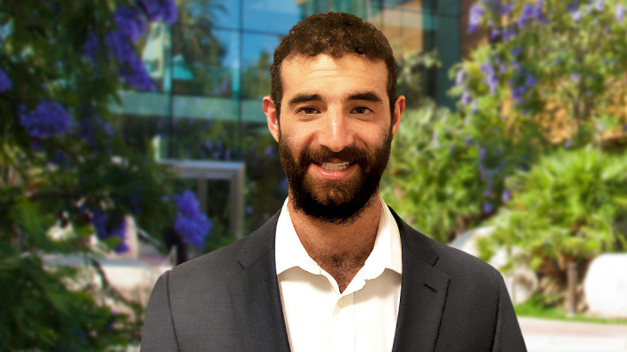 Portrait of Zach Jaffe smiling in a gray and white suit with a sunlit glass building surrounded by lush foliage in the background