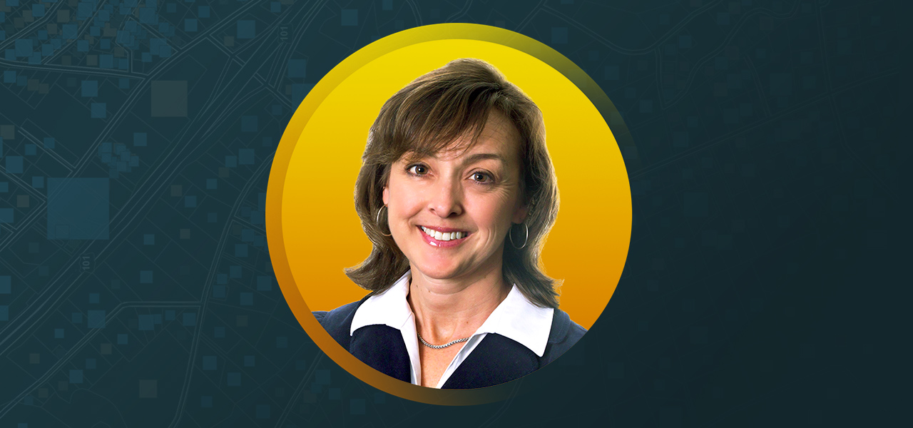 Donna Huey wearing a white dress shirt under a blue jacket and smiling with an abstract blue and yellow background