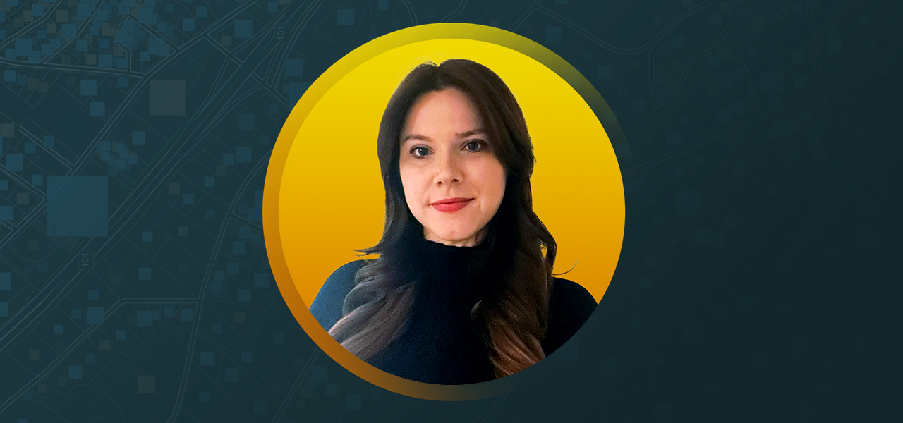 Portrait of Sophie Stouki smiling in a dark blue turtleneck with a flat golden yellow background in a round border
