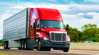 A red big rig truck moving down a greenery-lined highway under an overcast blue sky
