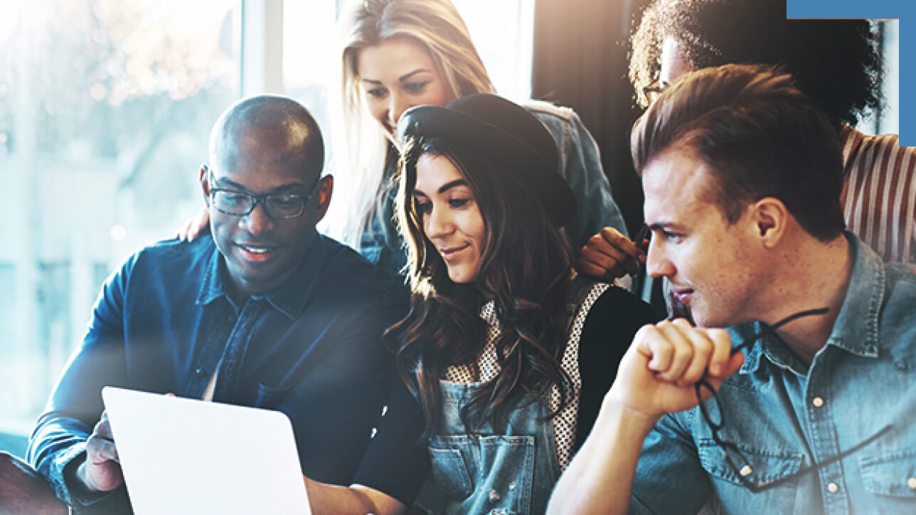 A group of five casually dressed people grouped closely around a shared laptop display in a sunlit office