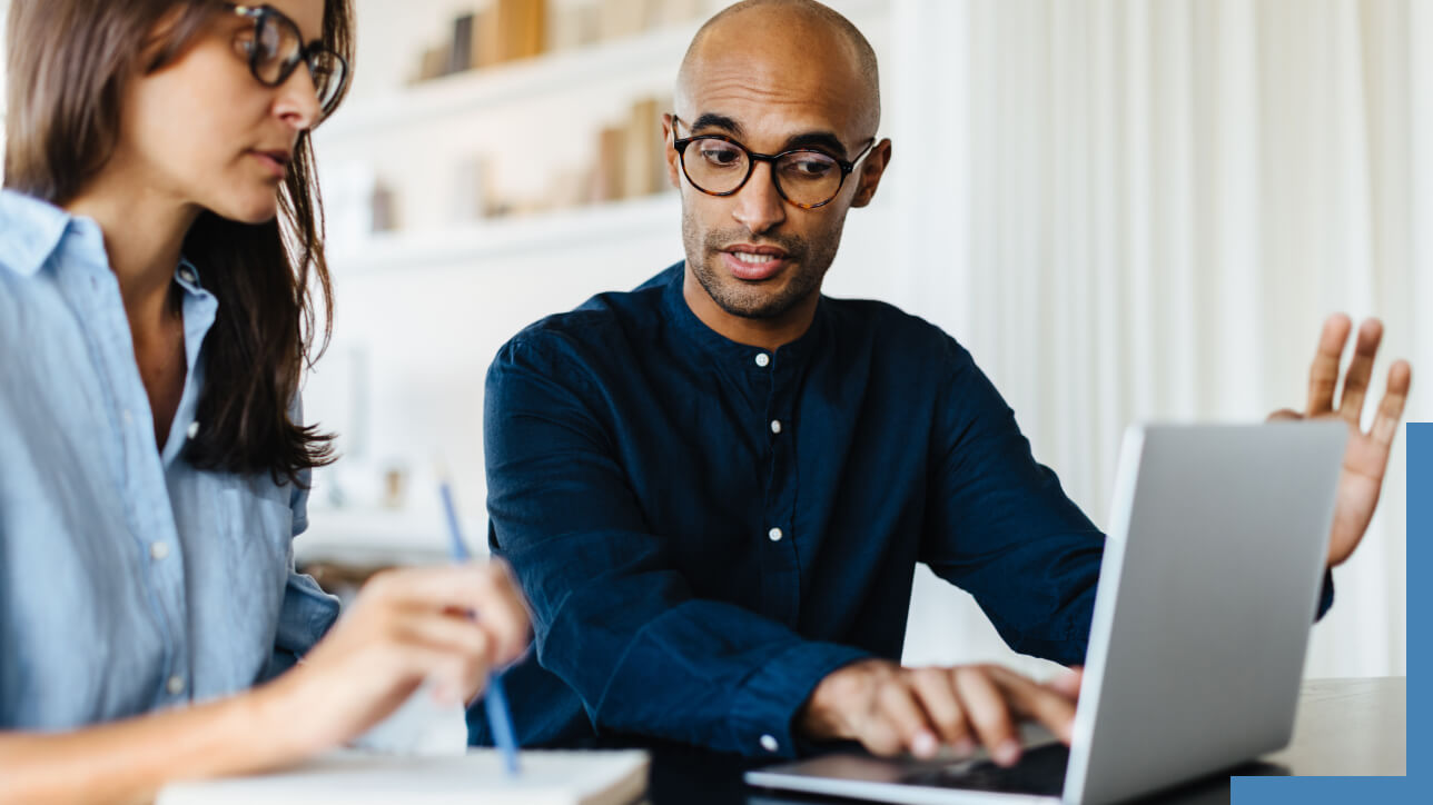 Two business professionals sharing a desk and discussing a laptop display in a brightly lit modern office