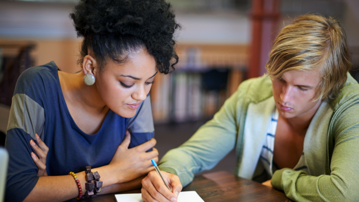 Two casually-dressed people lean intently over a wooden classroom desk, looking at the same paper document