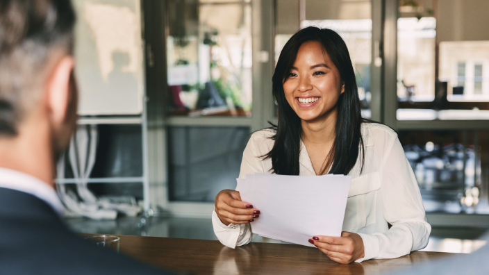 Two professionally-dressed people smile as they sit on opposite sides of an office desk, one holding a resume, in a well-lit modern office