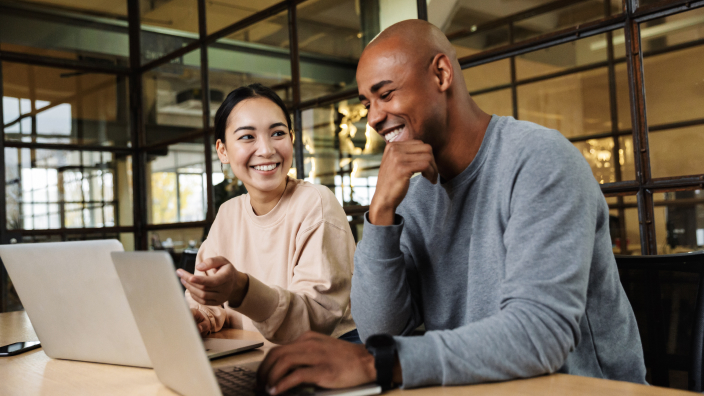 Two people wearing neutral sweaters laugh as they sit together at a conference table in a modern warmly-lit office, discussing two laptop displays