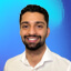 A portrait of Haseeb Malik smiling in a white collared shirt with an abstract white background dotted with blue and pink circles