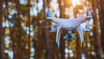 A closeup photo of a large white drone in mid-flight with a thick stand of trees in the unfocused background