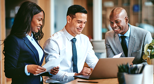 A photo of three smiling, professionally-dressed people, grouped around a laptop in a brightly-lit office environment