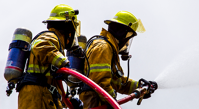 A photo of two firefighters in yellow hazmat suits, helmets, and gas masks, pointing a red fire hose at an off-camera target