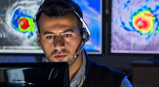 A photo of a man in a headset watching a computer monitor, while large screens behind him display aerial storm pattern maps