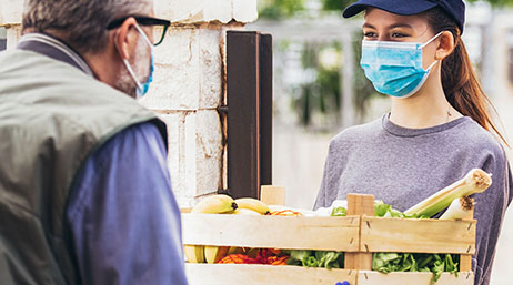 A photo of two casually dressed people standing outdoors wearing face masks as one hands the other a crate of colorful produce