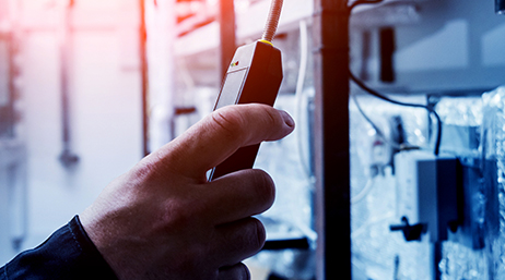 A closeup of a hand holding a methane gas detector near a wall of electrical equipment