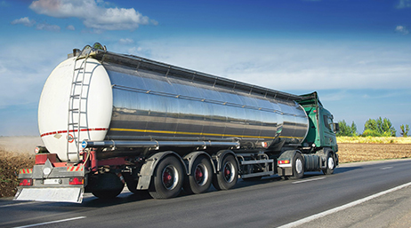 A silver and green big rig driving down a sunny highway through yellow croplands under a bright blue sky