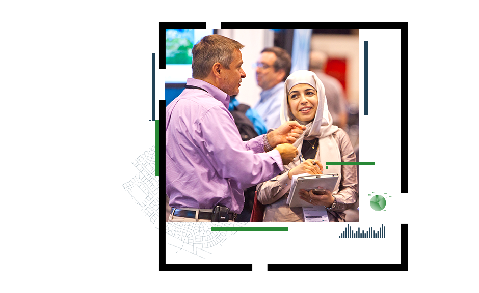 A conference attendee wearing a beige suit and headscarf holding a tablet in conversation with an exhibit staffer in a violet collared shirt