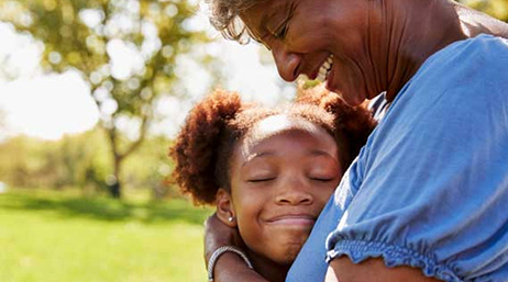 A photo of a smiling child embracing an adult wearing a casual blue shirt with a sunlit green field in the background