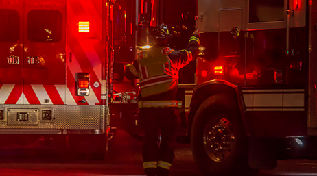 A photo of a first responder standing beside two fire engines on a street at night in a scene lit red from the trucks’ lights