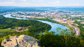 An aerial photo of a winding blue river bordered by green trees and a bustling city with hazy mountains in the distance