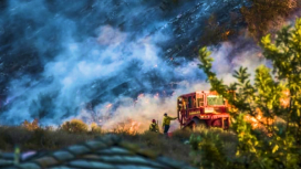 Fire personnel and a fire truck silhouetted against an orange wildfire with billowing black and gray smoke