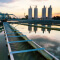 An industrial bridge over still gray water with a factory in the distance silhouetted against a hazy gold and white sunrise sky