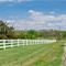 A long, paved, single-lane road near a white fence and surrounded by greenery