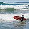 A child standing in the surf on a sunny beach wearing a blue wetsuit and carrying a red surfboard as he watches incoming waves