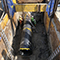 An infrastructure repair site with three people in hard hats working on a massive black pipe in a deep ditch