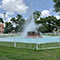 A large blue fountain sculpture surrounded by a white fence in a green tree-lined city park under a cloud-swept blue sky