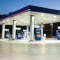 A clean, modern blue and white gas station with trees in the background silhouetted against a deep blue and red sunset sky