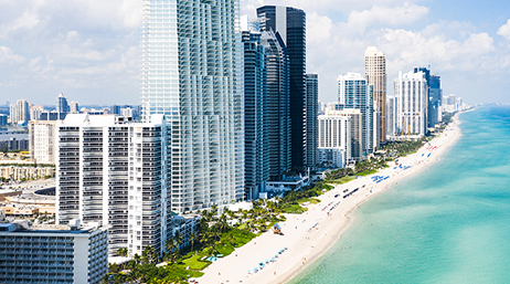 The beach coastline of Miami Beach, Florida with tall skyscrapers and people out on the white sand beaches next to the clear blue water