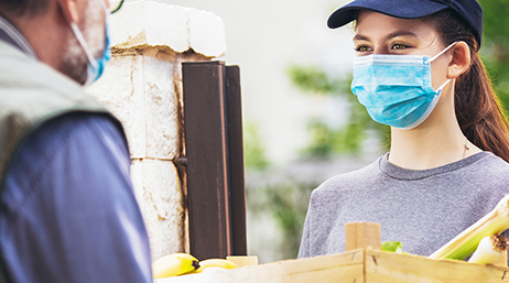 A person in a baseball cap and a mask handing a box of bananas and other food to someone else