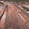 An aerial view of distant human figures examining muddy stretches of collapsed earth in a wide field of sandy scrub beside a railroad track